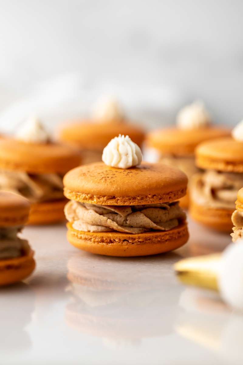 Pumpkin pie flavored French macarons recipe, complete with pumpkin pie frosting, pie crust center, and a dollop vanilla frosting on top! The perfect one bite dessert for fall and Thanksgiving menus. Dairy free
