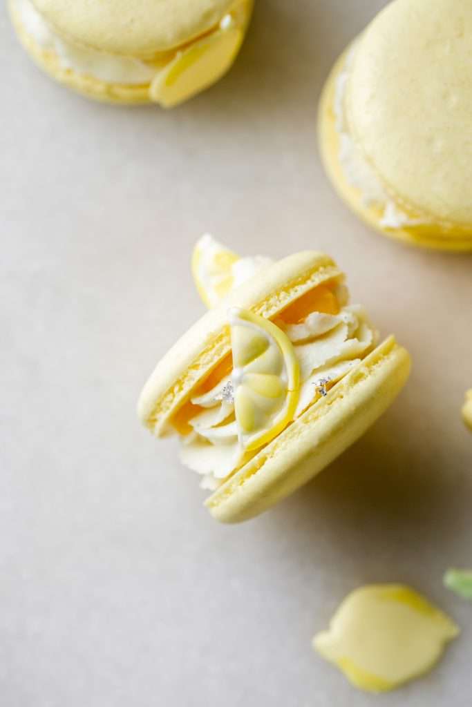 Lemon flavored French macarons filled with lemon vanilla bean frosting and lemon curd. Decorated with royal icing transfers. A dairy free and gluten free macaron recipe perfect for spring and summer.