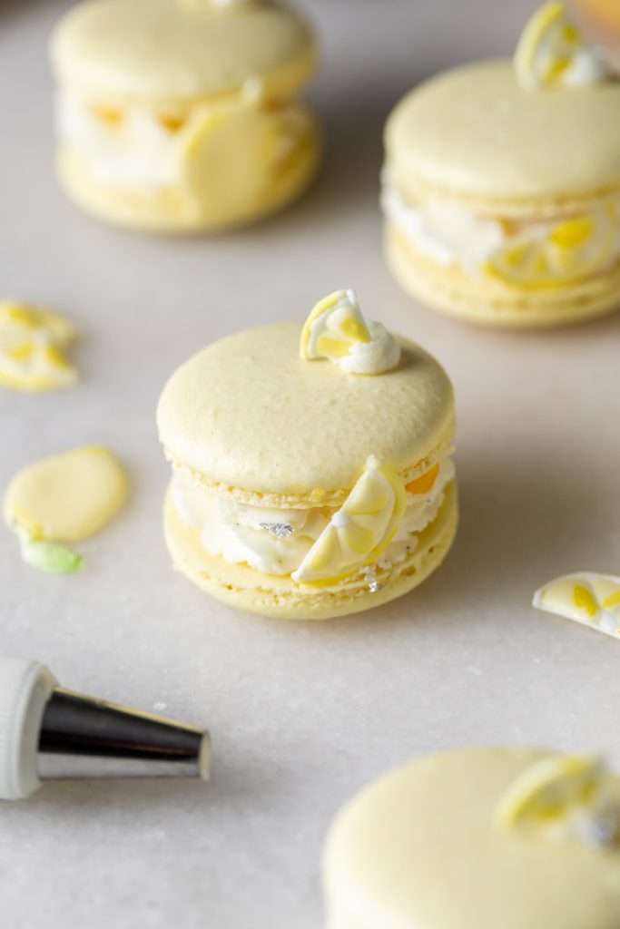 Lemon flavored French macarons filled with lemon vanilla bean frosting and lemon curd. Decorated with royal icing transfers. A dairy free and gluten free macaron recipe perfect for spring and summer.
