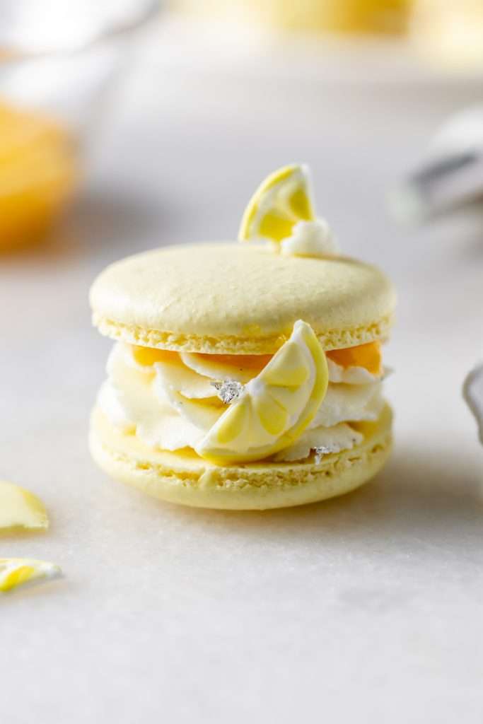 Lemon flavored French macarons filled with lemon vanilla bean frosting and lemon curd. Decorated with royal icing transfers. A dairy free and gluten free macaron recipe perfect for spring and summer.
