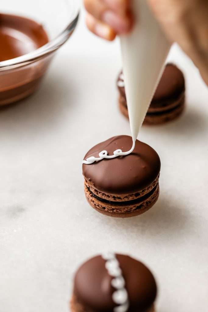 Hostess cupcake inspired macarons, with rich dark chocolate frosting and fluffy marshmallow filling. The shells are dipped in dark chocolate and decorated with icing loops to complete the look and taste! Check out the recipe, video and step by step instructions. Dairy free and gluten free option.