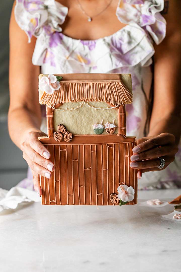 Large 10 inch tiki hut statement sugar cookie decorated with realist royal icing, featuring a thatched roof, bamboo details, and tropical accents and part of a pineapple coconut sugar cookie collection for a summer party.