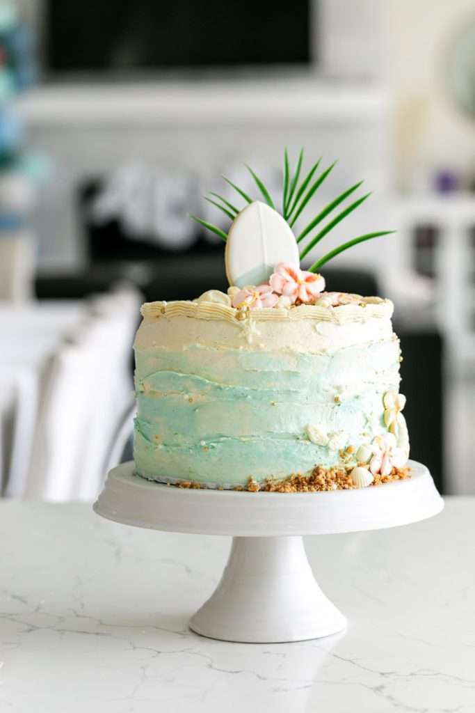 Tropical birthday cake decorated with palm leaves, hibiscus flowers, and beach inspired details.